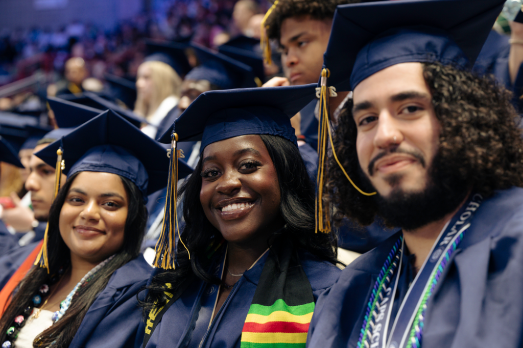 Three graduating students smile at the camera while wearing their regalia.